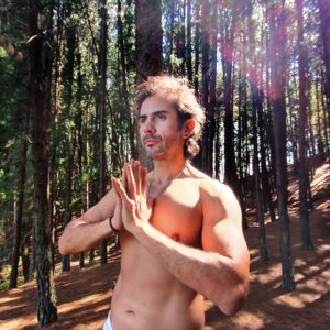 La serenidad en la naturaleza: Una persona sin camisa medita con las manos juntas frente al pecho en medio de un bosque, encontrando paz y conexión.